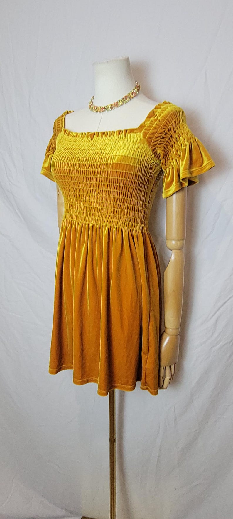 May include: Mustard yellow velvet dress with a square neckline and short, ruffled sleeves. The bodice is smocked, and the skirt is flared. A multi-colored beaded necklace is around the mannequin's neck. The dress is displayed on a wooden mannequin.