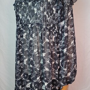 Puede incluir: Vestido con estampado floral en blanco y negro con mangas largas y dobladillo con volantes. El vestido presenta un cuello redondo y un corpi&ntilde;o plisado. La tela parece ligera y semitransparente, con un delicado estampado floral.
