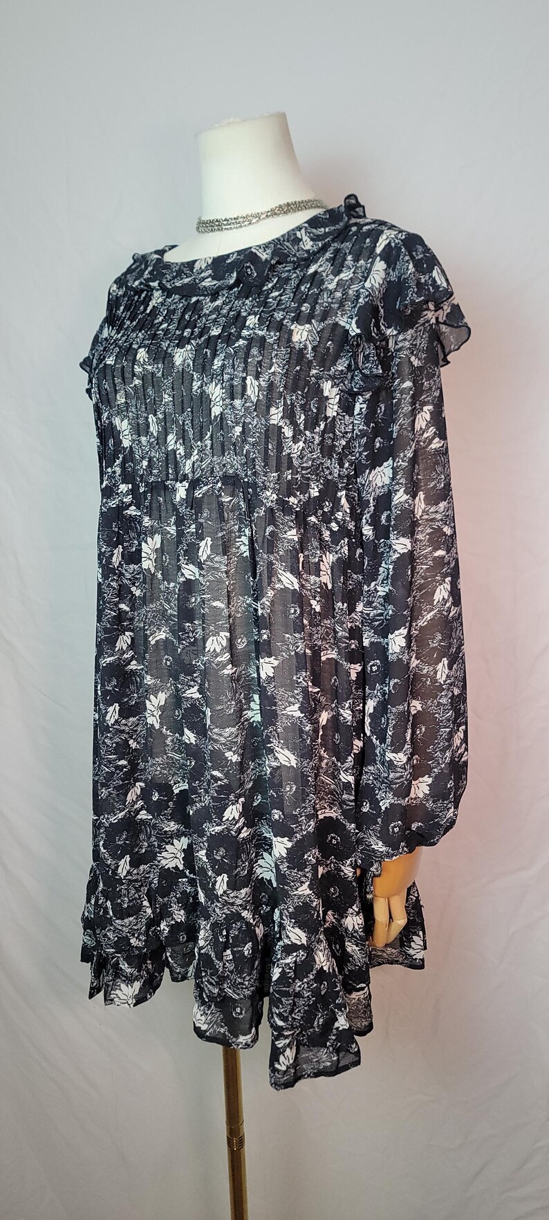 Puede incluir: Vestido con estampado floral en blanco y negro con mangas largas y dobladillo con volantes. El vestido presenta un cuello redondo y un corpi&ntilde;o plisado. La tela parece ligera y semitransparente, con un delicado estampado floral.