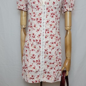 Puede incluir: Un minivestido blanco con un estampado floral rojo y mangas cortas abullonadas. El vestido tiene un escote coraz&oacute;n y una fila de peque&ntilde;os botones. Un bolso burdeos cuelga del brazo del maniqu&iacute;.