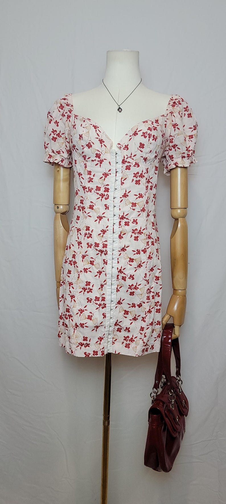 Puede incluir: Un minivestido blanco con un estampado floral rojo y mangas cortas abullonadas. El vestido tiene un escote coraz&oacute;n y una fila de peque&ntilde;os botones. Un bolso burdeos cuelga del brazo del maniqu&iacute;.