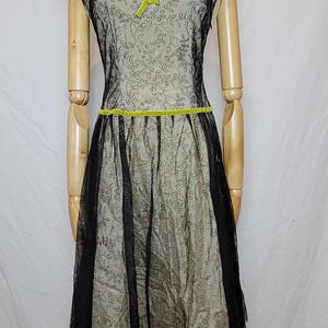 Pu&ograve; includere: Un abito in stile vintage su un manichino. L'abito presenta uno strato inferiore beige con un rivestimento nero a pois. Ha una gonna in tulle nero, maniche a cappuccio e un fiocco e una cintura verde lime. &Egrave; visibile anche una collana di perle.