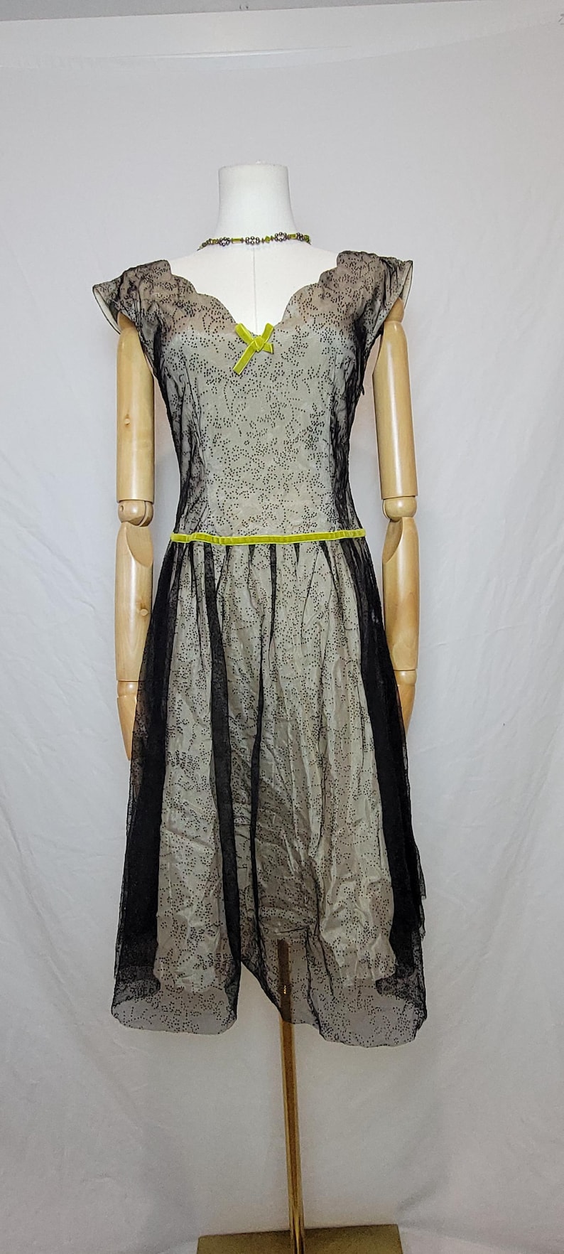 Pu&ograve; includere: Un abito in stile vintage su un manichino. L'abito presenta uno strato inferiore beige con un rivestimento nero a pois. Ha una gonna in tulle nero, maniche a cappuccio e un fiocco e una cintura verde lime. &Egrave; visibile anche una collana di perle.