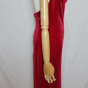 May include: A vibrant red, floor-length dress with a draped cowl neckline. The dress is sleeveless and has a fitted silhouette, showcasing a sleek and elegant design. The dress is displayed on a mannequin.