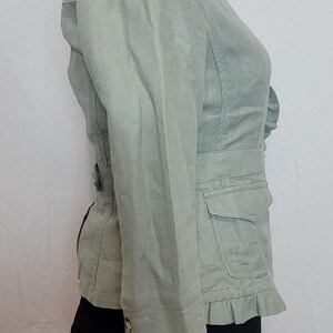 May include: A pale green jacket with a fitted waist, small pockets, and a frilled hem. The jacket has long sleeves with button details and a notched collar. The jacket is displayed on a mannequin.