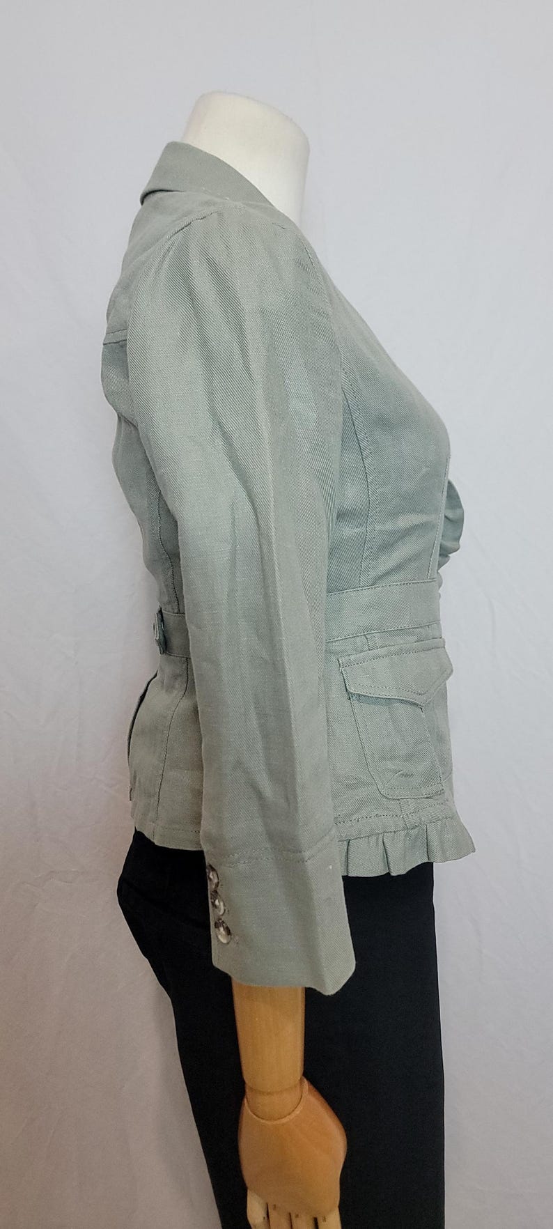 May include: A pale green jacket with a fitted waist, small pockets, and a frilled hem. The jacket has long sleeves with button details and a notched collar. The jacket is displayed on a mannequin.