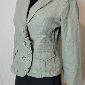 May include: A pale green blazer with a ruffled front, two button closure, and two front pockets. The blazer has three-quarter sleeves and is displayed on a mannequin. The mannequin is wearing a black top and black trousers.
