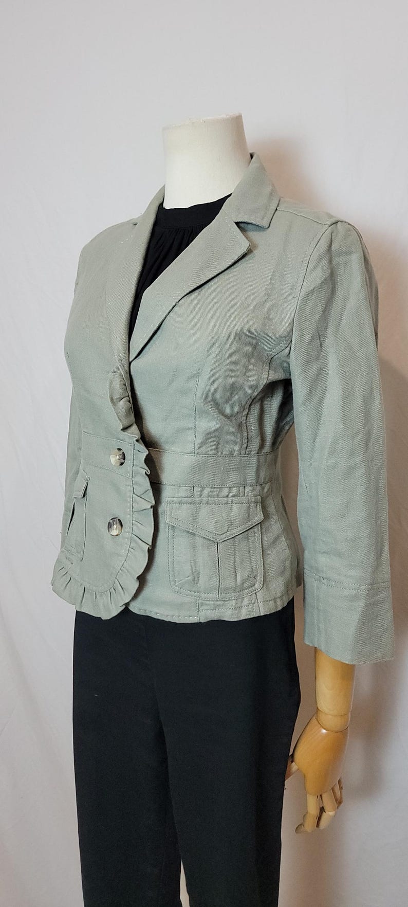 May include: A pale green blazer with a ruffled front, two button closure, and two front pockets. The blazer has three-quarter sleeves and is displayed on a mannequin. The mannequin is wearing a black top and black trousers.