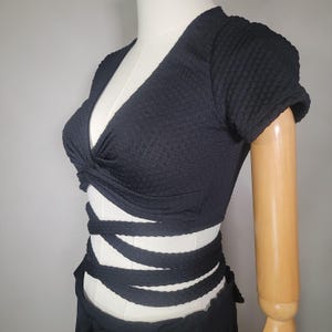 May include: Black crop top with a twist detail at the front and a tie closure at the waist. The top is made of a textured fabric.