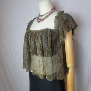 May include: A gold and black pleated top with a square neckline and ruffled sleeves is displayed on a mannequin. A layered necklace with brown and black beads is around the mannequin's neck. The bottom of the mannequin is wearing a black skirt.