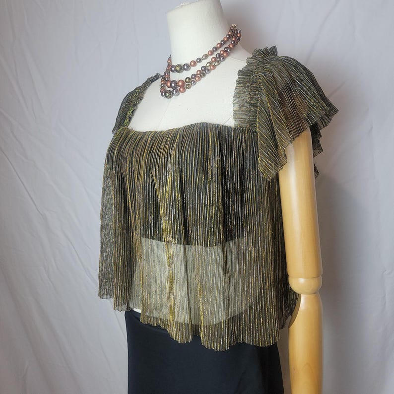 May include: A gold and black pleated top with a square neckline and ruffled sleeves is displayed on a mannequin. A layered necklace with brown and black beads is around the mannequin's neck. The bottom of the mannequin is wearing a black skirt.