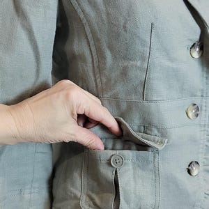 May include: A sage green blazer with a notched lapel, two front pockets, and decorative buttons. The blazer has a ruffled hem and is made of a textured fabric.