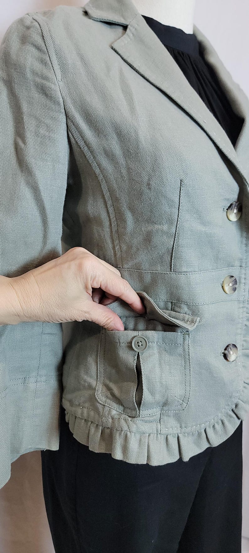 May include: A sage green blazer with a notched lapel, two front pockets, and decorative buttons. The blazer has a ruffled hem and is made of a textured fabric.