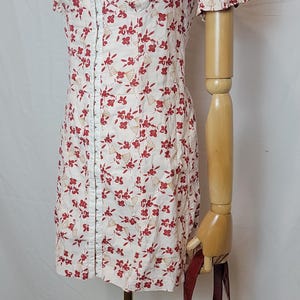 Puede incluir: Un minivestido blanco con estampado floral rojo, mangas cortas abullonadas y escote en V. El vestido se exhibe en un maniqu&iacute; con un bolso de mano rojo. Tambi&eacute;n se ve un collar plateado.