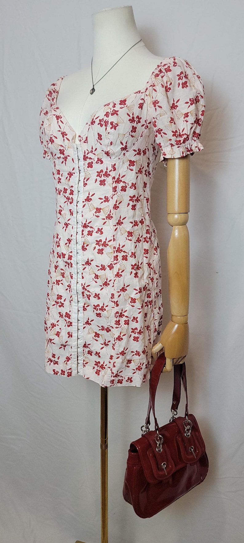 Puede incluir: Un minivestido blanco con estampado floral rojo, mangas cortas abullonadas y escote en V. El vestido se exhibe en un maniqu&iacute; con un bolso de mano rojo. Tambi&eacute;n se ve un collar plateado.