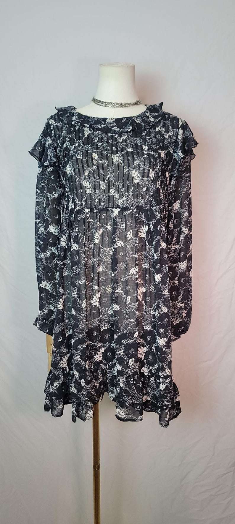 Puede incluir: Vestido con estampado floral en blanco y negro con mangas largas y cuello con volantes. El vestido se exhibe en un maniqu&iacute;. El vestido tiene un volante escalonado en el dobladillo y un delicado collar plateado.