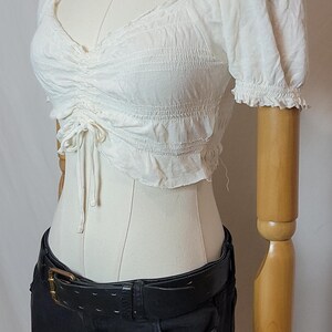 Op de afbeelding: Een witte crop top met korte mouwen, een gerimpeld lijfje en een trekkoord, gecombineerd met een zwarte jeans en een zwarte riem. Een zilveren ketting om de nek van de mannequin. De top heeft een V-hals en gefranjerde mouwen.