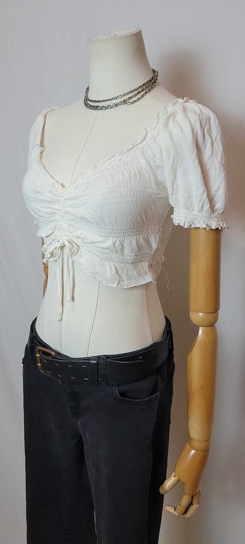 Op de afbeelding: Een witte crop top met korte mouwen, een gerimpeld lijfje en een trekkoord, gecombineerd met een zwarte jeans en een zwarte riem. Een zilveren ketting om de nek van de mannequin. De top heeft een V-hals en gefranjerde mouwen.