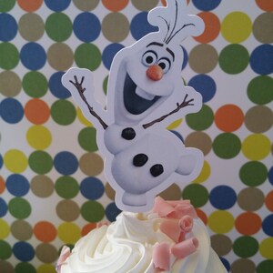 May include: A cupcake with white frosting and pink chocolate shavings, topped with a cutout of the character Olaf from the movie Frozen. The background features a pattern of colorful polka dots.
