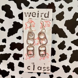 Op de afbeelding: Een paar zilveren oorbellen gemaakt van gerecyclede blikjes. De oorbellen hangen aan een witte kaart met de tekst "weird class" erop.