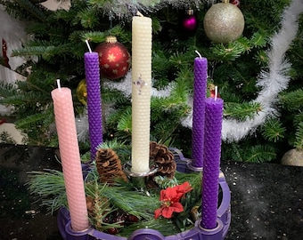 May include: An Advent wreath featuring a purple circular base with a nativity scene relief, holding four tall honeycomb-textured candles in pink, purple, and white. The white candle has a small cross embellishment. Pine branches, pinecones, and red poinsettia accents fill the center.