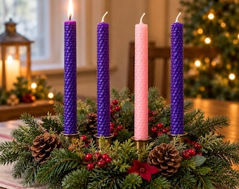 May include: An Advent wreath featuring three purple and one pink textured beeswax taper candles in brass holders, surrounded by evergreen boughs, pinecones, and red berries. The first purple candle is lit with a visible flame.