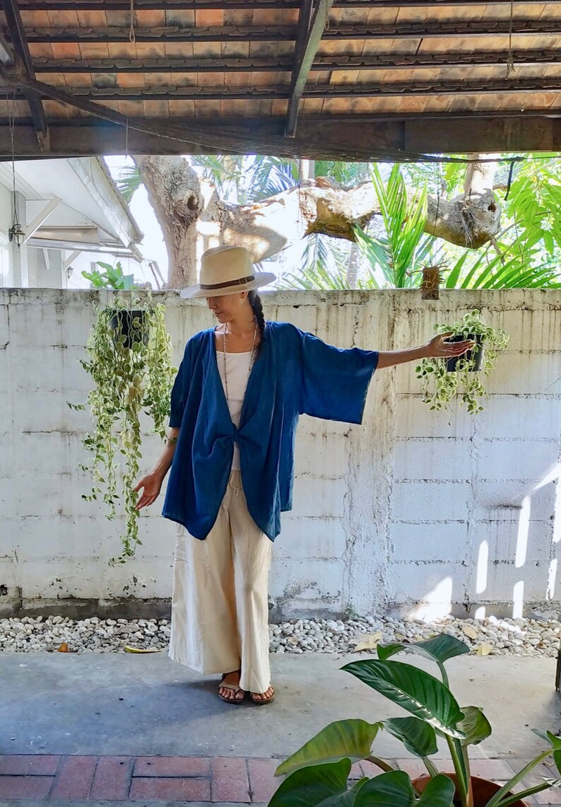Puede incluir: Una mujer con un kimono de lino azul con un nudo en la parte delantera, un sombrero blanco y pantalones blancos. Est&aacute; de pie frente a una pared de ladrillo blanco con plantas colgando.