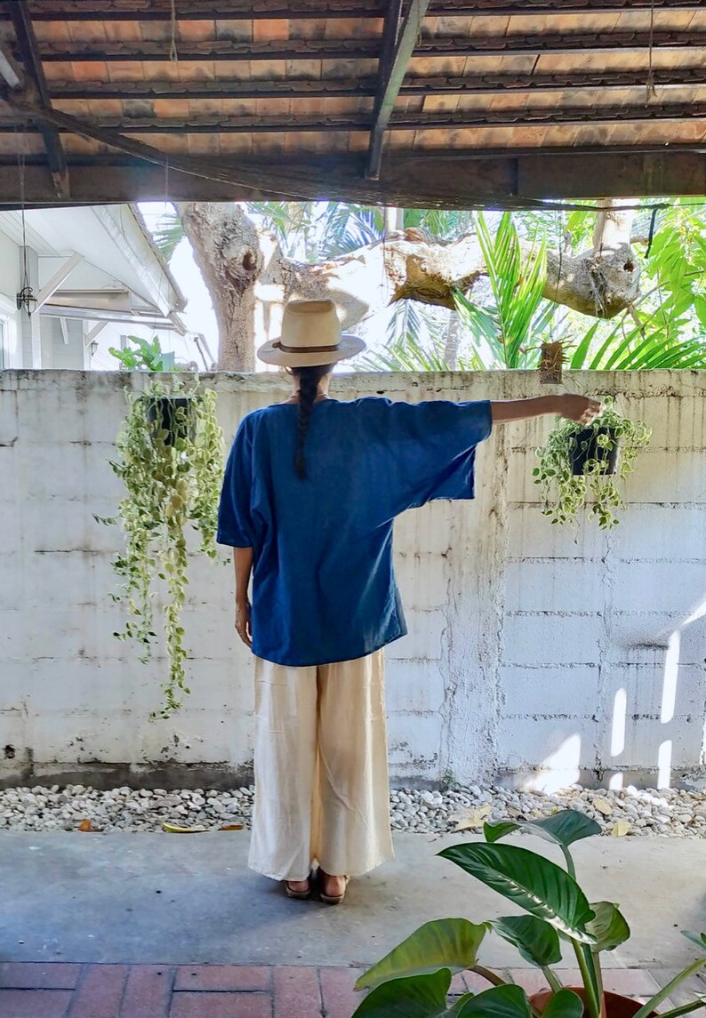 Puede incluir: Una persona con una camisa azul y pantalones beige se encuentra frente a una pared de ladrillo blanco. Lleva un sombrero de paja y est&aacute; de espaldas a la c&aacute;mara. Hay plantas colgantes en la pared.