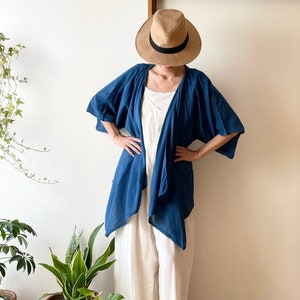 Puede incluir: Una mujer lleva un kimono de lino azul sobre un vestido de lino blanco y un sombrero de paja de ala ancha. Est&aacute; de pie frente a una pared blanca con plantas en macetas en el suelo.