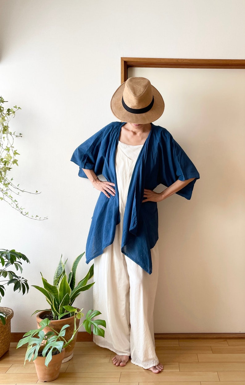 Puede incluir: Una mujer lleva un kimono de lino azul sobre un vestido de lino blanco y un sombrero de paja de ala ancha. Est&aacute; de pie frente a una pared blanca con plantas en macetas en el suelo.