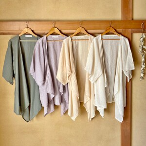 Puede incluir: Cuatro c&aacute;rdigans de lino ligero en diferentes colores colgados en perchas de madera contra una pared beige. Los c&aacute;rdigans son en tonos de verde, morado, beige y blanco.