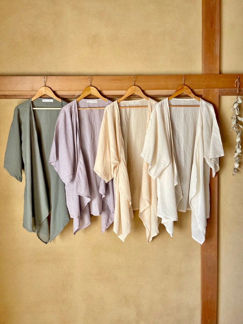 Puede incluir: Cuatro c&aacute;rdigans de lino ligero en diferentes colores colgados en perchas de madera contra una pared beige. Los c&aacute;rdigans son en tonos de verde, morado, beige y blanco.
