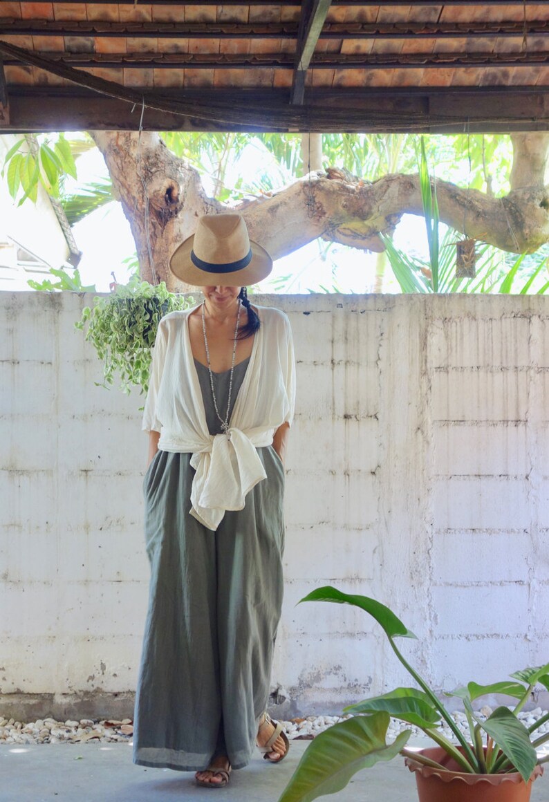 Puede incluir: Una mujer con un sombrero de paja de ala ancha, una chaqueta de lino blanca y pantalones de lino verde holgados. Est&aacute; de pie frente a una pared blanca con una planta en maceta en primer plano.