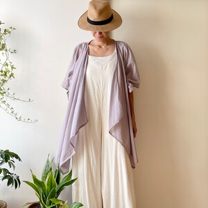 Puede incluir: Una mujer con un mono blanco, una chaqueta de punto de color lila claro y un sombrero de paja de ala ancha.