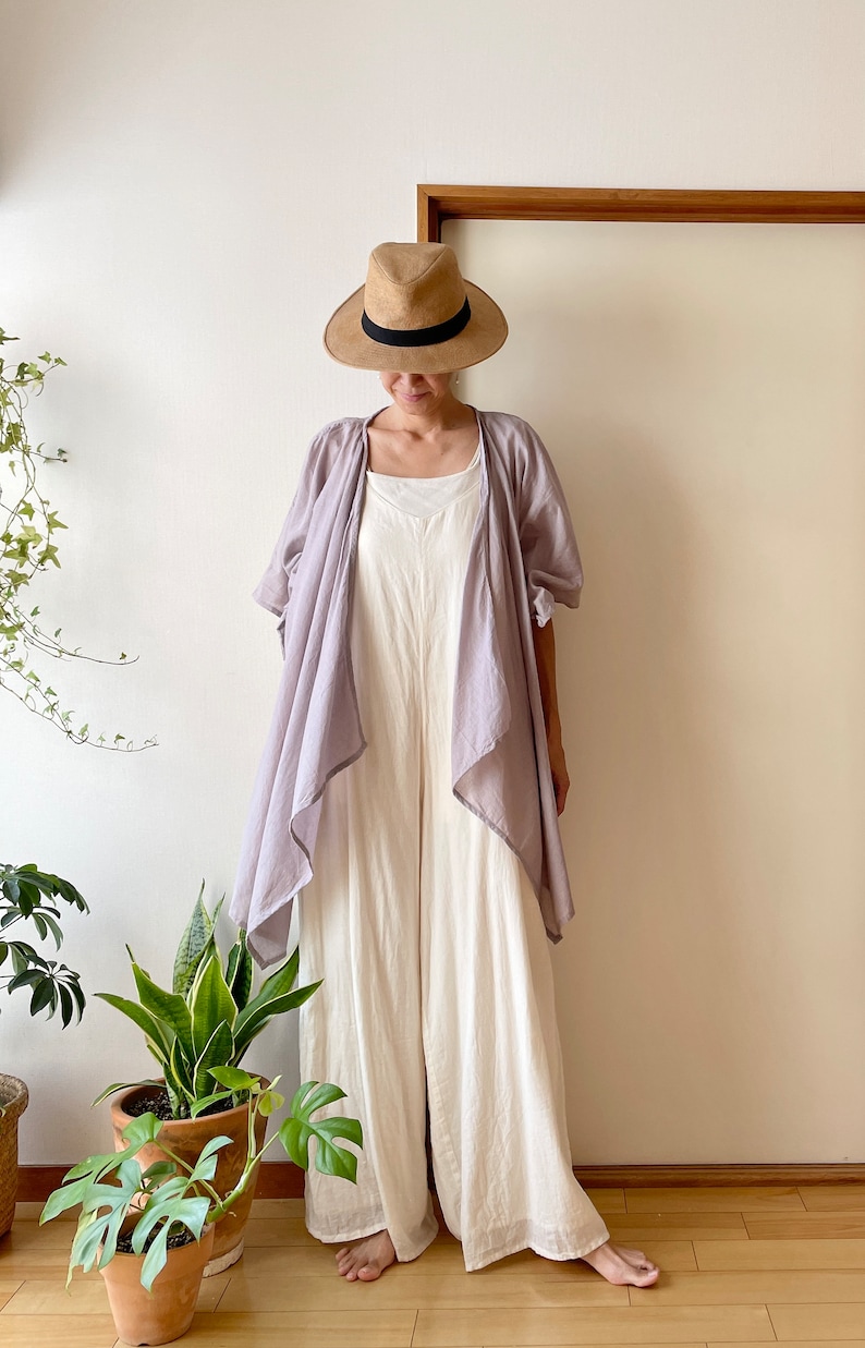 Puede incluir: Una mujer con un mono blanco, una chaqueta de punto de color lila claro y un sombrero de paja de ala ancha.