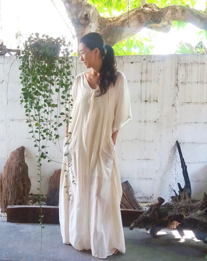Puede incluir: Una mujer lleva un vestido largo, blanco y fluido, de corte holgado y escote redondo. Est&aacute; de pie frente a una pared blanca con una planta verde colgando en el fondo.