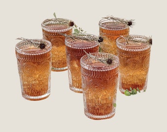 May include: A set of five clear glass tumblers featuring a textured, embossed pattern of circular medallions and vertical ridges. Each glass is filled with an amber-colored liquid and garnished with a sprig of rosemary and a small pinecone.