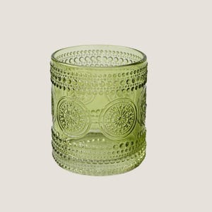 Puede incluir: Un vaso de vidrio verde con un patrón decorativo de círculos y puntos. El vaso tiene forma cilíndrica y una superficie texturizada. El diseño se repite alrededor de la circunferencia del vaso.