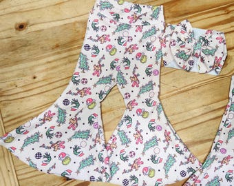 May include: White baby pants with a flared leg and matching headband. The pants and headband feature a repeating pattern of Christmas-themed illustrations in pink, green, yellow, and red. The illustrations include Christmas trees, snowmen, and Santa Claus.