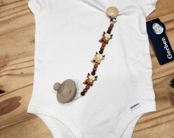 Könnte beinhalten: Ein weißer Baby-Strampler mit einem braunen und weißen Holz-Schnullerclip. Der Schnullerclip hat einen Holzclip und zeigt ein Muster aus kleinen braunen Kühen.