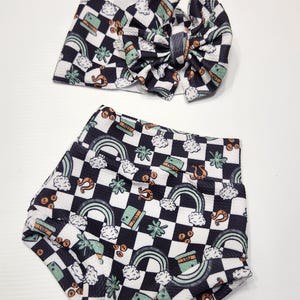 May include: A baby headband and shorts set with a black and white chequered pattern. The pattern includes rainbows, shamrocks, pots of gold, and horseshoes. The headband has a large bow.