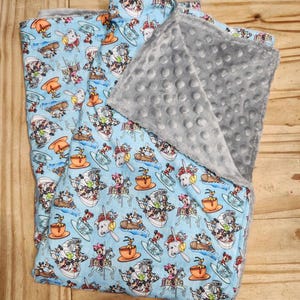 Puede incluir: Una manta para bebé azul y gris con un estampado de personajes de Disney. La manta presenta a Mickey Mouse, Minnie Mouse, Donald Duck, Goofy y Pluto. El estampado está sobre un fondo azul claro con detalles blancos. La parte posterior de la manta es de un gris suave.