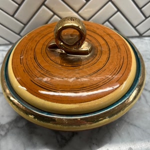 May include: A round ceramic lidded dish with a gold-colored handle. The lid has a swirling pattern in brown and black on a cream background. The dish has a gold rim and a blue band.
