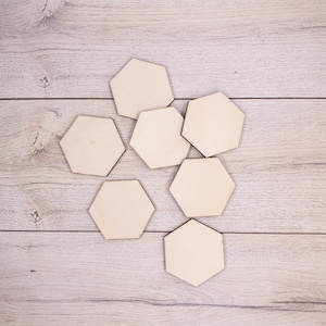 Puede incluir: Siete posavasos hexagonales de madera sin terminar, dispuestos sobre una superficie clara con vetas de madera. Los posavasos son de madera natural, listos para personalizar. Cada posavasos mide unos 10 cm de diámetro.