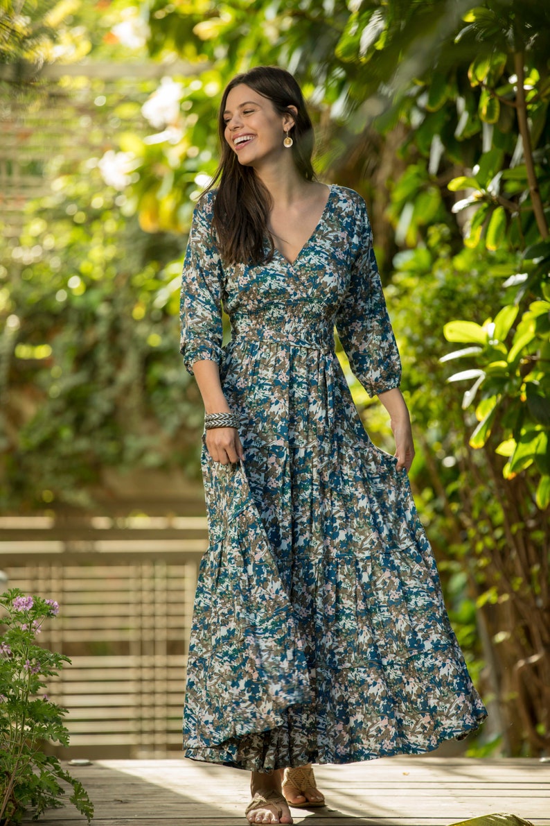 May include: A woman wearing a long, blue and green floral print dress with a v-neck and 3/4 sleeves. She is standing in a garden setting.
