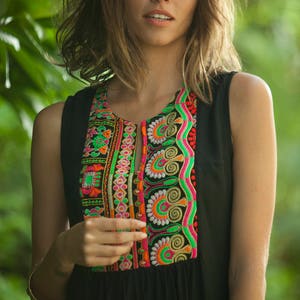 May include: A woman wearing a black sleeveless dress with colorful embroidery around the neckline. The embroidery features a variety of patterns and colors, including pink, green, orange, and white.
