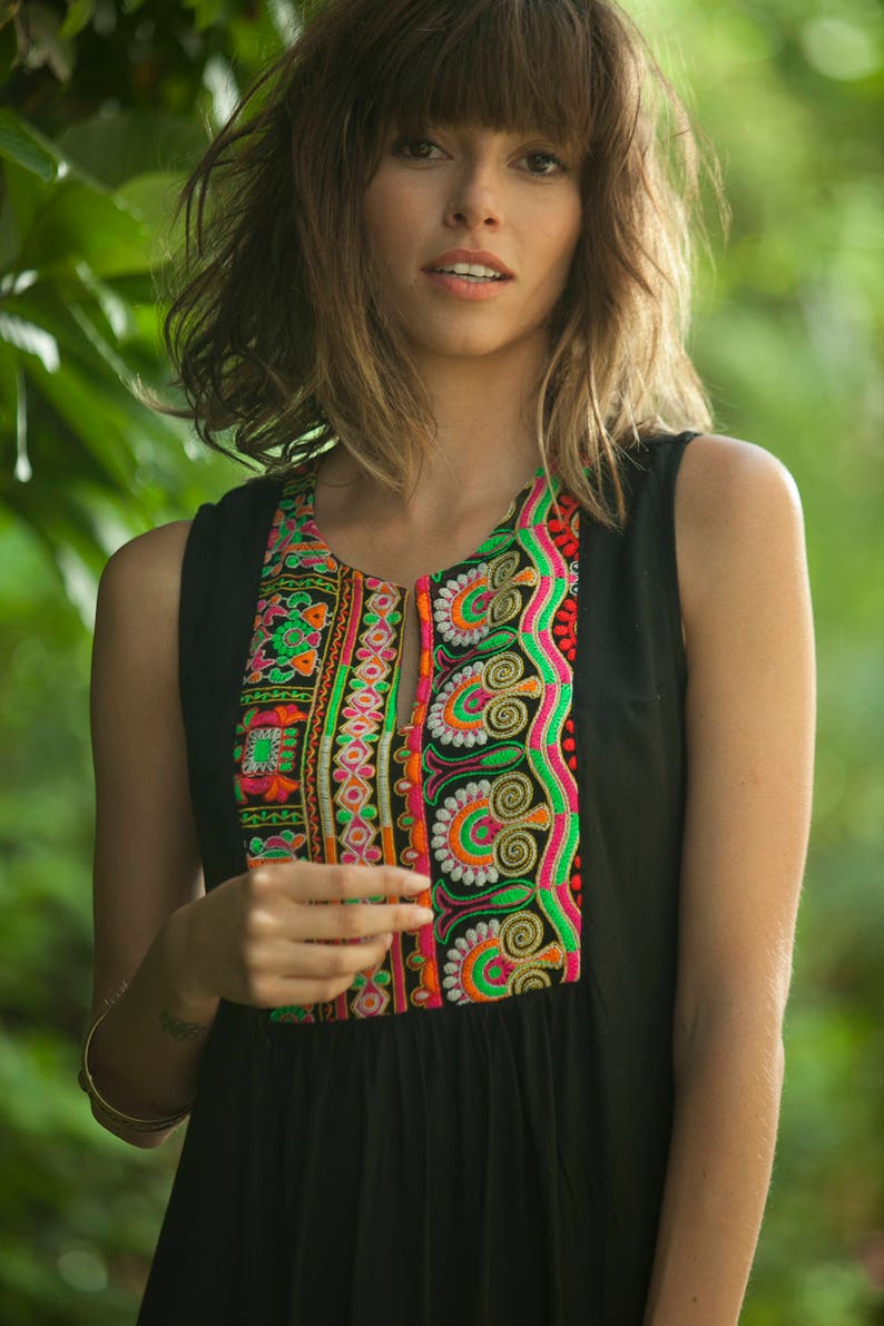 May include: A woman wearing a black sleeveless dress with colorful embroidery around the neckline. The embroidery features a variety of patterns and colors, including pink, green, orange, and white.