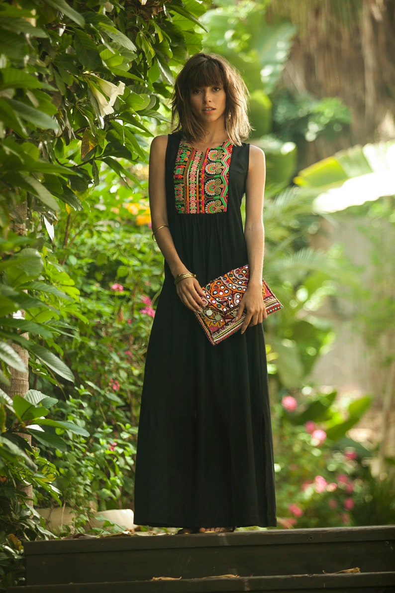 May include: A woman wearing a black maxi dress with colorful embroidery on the bodice. She is holding a colorful clutch purse with mirror accents.