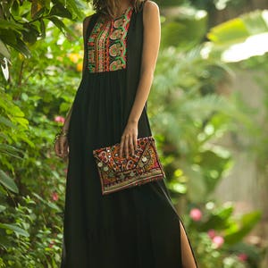 May include: A woman wearing a black maxi dress with colorful embroidery on the neckline. She is holding a colorful embroidered clutch bag with silver accents.