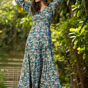 May include: A woman wearing a long, flowing blue and white floral dress with a cinched waist and a tie belt. She is standing on a wooden deck with green foliage in the background.